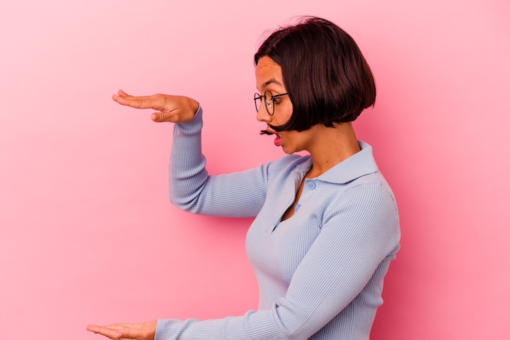 Foto mujer joven de raza mixta aislada sobre fondo rosa conmocionado y asombrado sosteniendo un espacio de copia entre las manos.