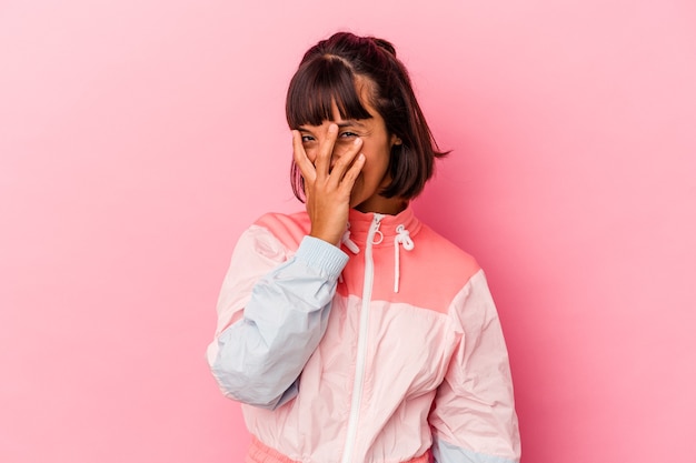 Mujer joven de raza mixta aislada sobre fondo rosa parpadear a la cámara a través de los dedos, avergonzado cubriendo la cara.