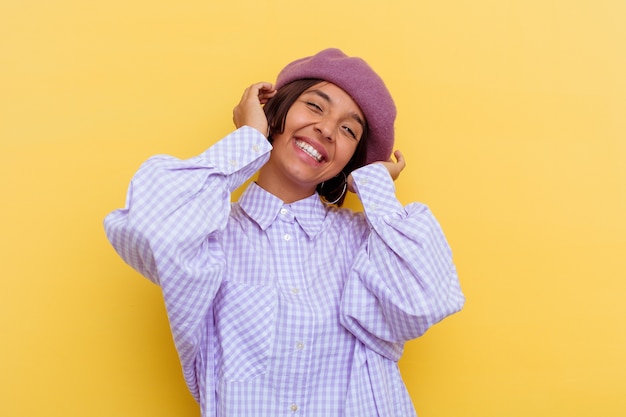 Mujer joven de raza mixta con una boina aislada sobre fondo amarillo se ríe con alegría manteniendo las manos en la cabeza. Concepto de felicidad.