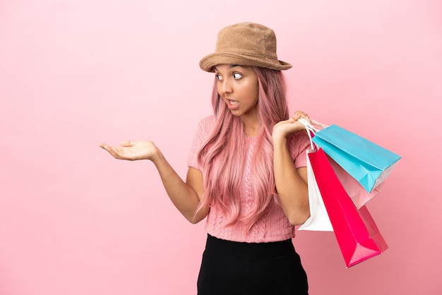 Mujer joven de raza mixta con bolsa de compras aislada sobre fondo rosa con expresión de sorpresa mientras mira de lado