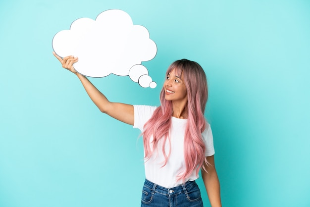 Mujer joven de raza mixta con cabello rosado aislado sobre fondo azul sosteniendo un bocadillo de diálogo de pensamiento