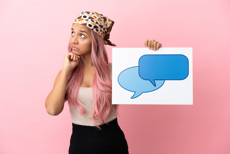 Foto mujer joven de raza mixta con cabello rosado aislado sobre fondo rosa sosteniendo un cartel con el icono de la burbuja del discurso y el pensamiento