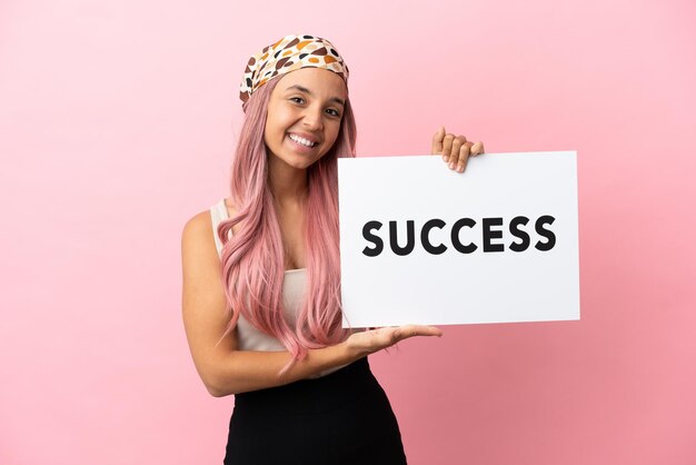 Mujer joven de raza mixta con cabello rosado aislado sobre fondo rosa sosteniendo un cartel con el texto ÉXITO con expresión feliz