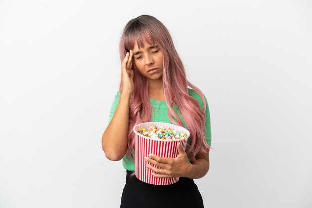 Mujer joven de raza mixta con cabello rosado comiendo palomitas de maíz aislado sobre fondo blanco con dolor de cabeza