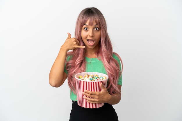 Mujer joven de raza mixta con cabello rosado comiendo palomitas de maíz aislado sobre fondo blanco haciendo gesto de teléfono. Llámame señal