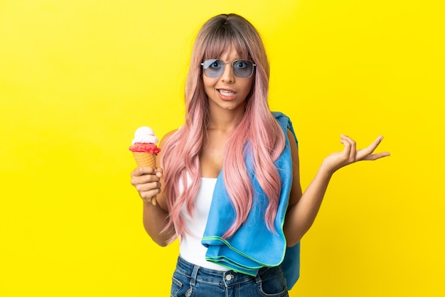 Mujer joven de raza mixta con cabello rosado con helado aislado sobre fondo amarillo con expresión facial sorprendida