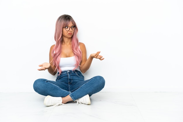 Mujer joven de raza mixta con cabello rosado sentada en el suelo aislado sobre fondo blanco con expresión facial sorpresa