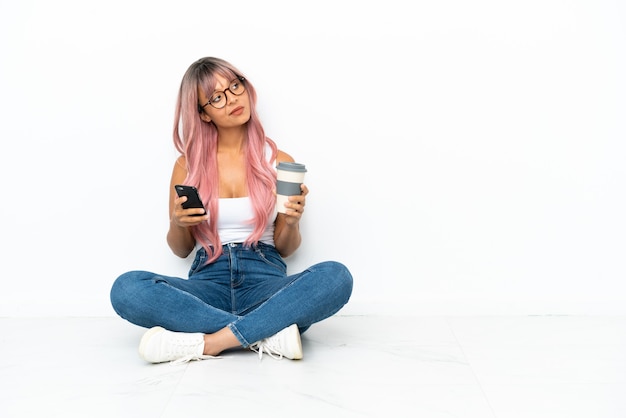 Mujer joven de raza mixta con cabello rosado sentada en el suelo aislado sobre fondo blanco sosteniendo un café para llevar y un móvil mientras piensa en algo