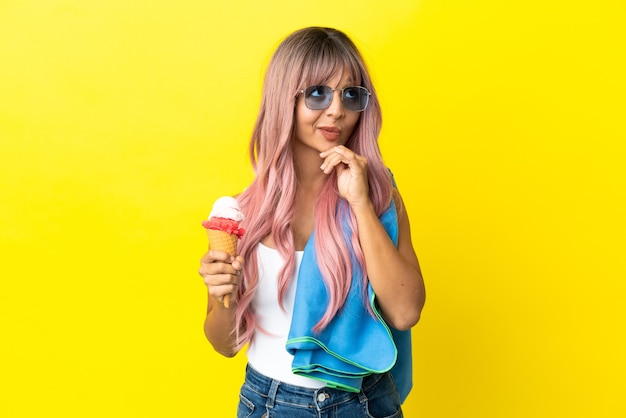 Mujer joven de raza mixta con cabello rosado sosteniendo helado aislado sobre fondo amarillo y mirando hacia arriba