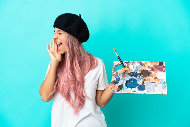 Mujer joven de raza mixta con cabello rosado sosteniendo una paleta aislada sobre fondo azul gritando con la boca abierta hacia el lado