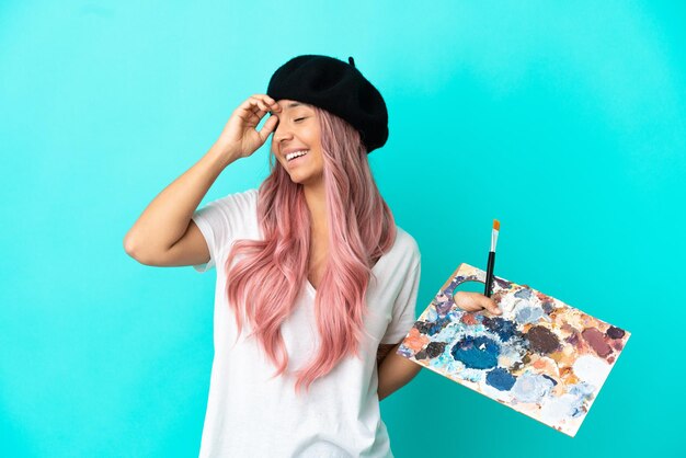 Mujer joven de raza mixta con cabello rosado sosteniendo una paleta aislada sobre fondo azul sonriendo mucho