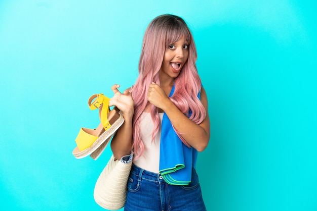 Mujer joven de raza mixta con cabello rosado sosteniendo sandalias de verano aisladas sobre fondo azul celebrando una victoria