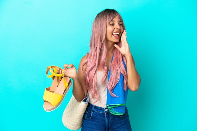 Mujer joven de raza mixta con cabello rosado sosteniendo sandalias de verano aisladas sobre fondo azul gritando con la boca abierta hacia el lado