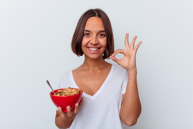 Mujer joven de raza mixta comiendo cereales aislados en la pared blanca alegre y confiado mostrando gesto ok.