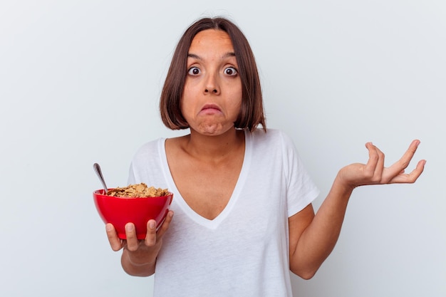 Mujer joven de raza mixta comiendo cereales aislados en la pared blanca se encoge de hombros y abre los ojos confundidos.