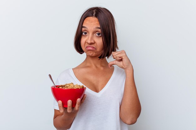 Mujer joven de raza mixta comiendo cereales aislados en la pared blanca se siente orgullosa y segura de sí misma, ejemplo a seguir.