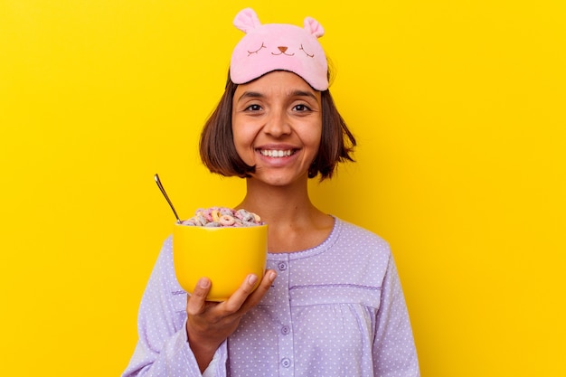 Mujer joven de raza mixta comiendo cereales vistiendo un pijama aislado sobre fondo amarillo feliz, sonriente y alegre.