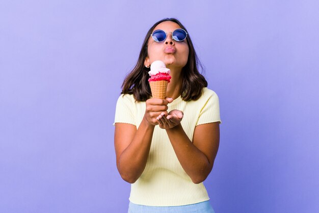 Mujer joven de raza mixta comiendo un helado doblando los labios y sosteniendo las palmas de las manos para enviar un beso de aire.