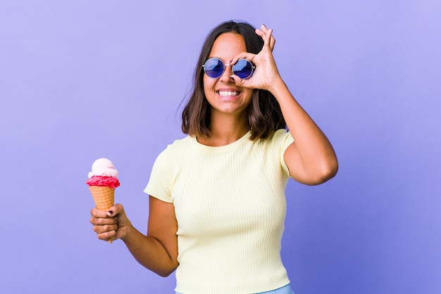 Mujer joven de raza mixta comiendo un helado emocionado manteniendo el gesto ok en el ojo.