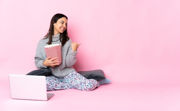 Mujer joven de raza mixta comiendo palomitas de maíz mientras ve una película en la computadora portátil apuntando hacia el lado para presentar un producto
