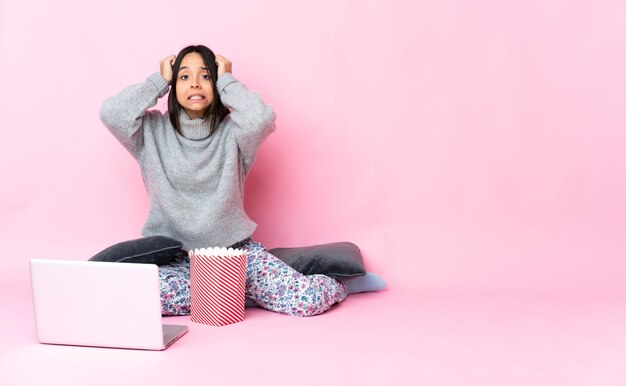 Mujer joven de raza mixta comiendo palomitas de maíz mientras ve una película en la computadora portátil haciendo gesto nervioso