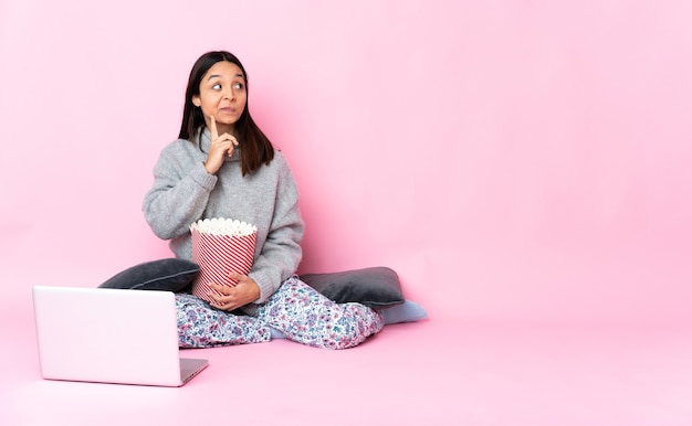 Mujer joven de raza mixta comiendo palomitas de maíz mientras ve una película en la computadora portátil que tiene dudas y pensamiento