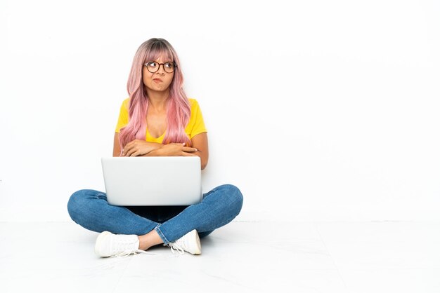 Mujer joven de raza mixta con una computadora portátil con el pelo rosa sentado en el suelo aislado sobre fondo blanco haciendo gesto de dudas mientras levanta los hombros