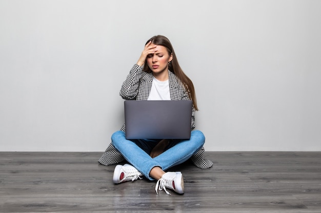 Mujer joven de raza mixta con una computadora portátil sentada en el suelo con expresión cansada y aburrida