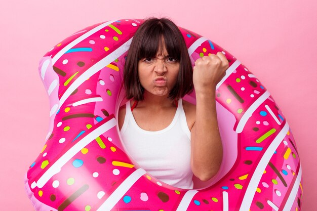 Mujer joven de raza mixta con donut inflable aislado sobre fondo rosa mostrando el puño a la cámara, expresión facial agresiva.