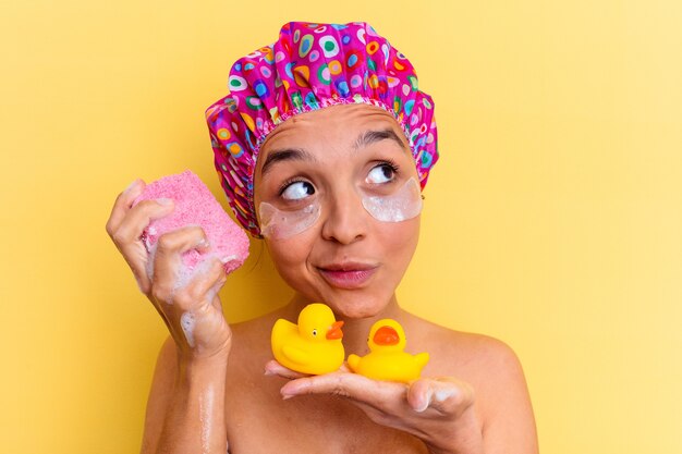 Mujer joven de raza mixta con gorro de ducha sosteniendo una esponja y patos de goma aislado sobre fondo amarillo