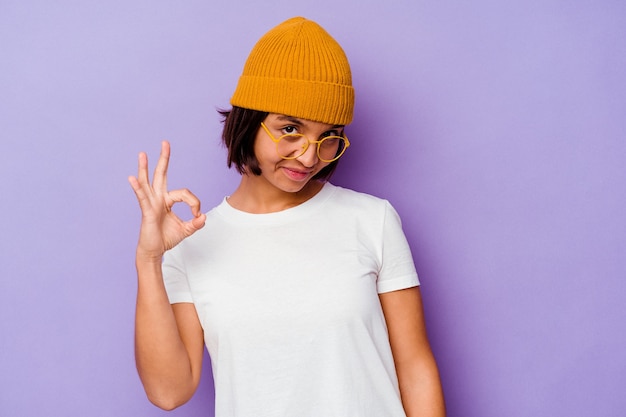 Mujer joven de raza mixta con un gorro de lana aislado sobre fondo púrpura alegre y confiado mostrando gesto ok.