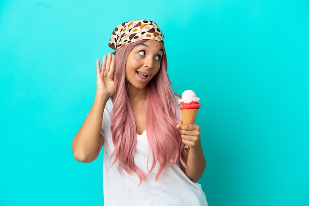 Mujer joven de raza mixta con un helado de cucurucho aislado sobre fondo azul escuchando algo poniendo la mano en la oreja