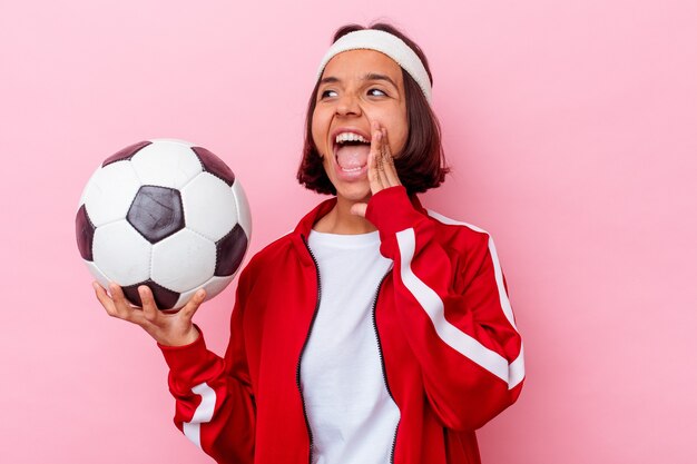 Mujer joven de raza mixta jugando al fútbol aislado en la pared rosa gritando y sosteniendo la palma cerca de la boca abierta.