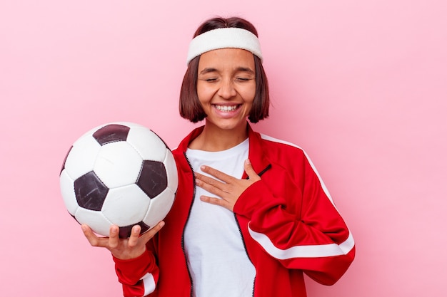 Mujer joven de raza mixta jugando al fútbol aislado sobre fondo rosa se ríe a carcajadas manteniendo la mano en el pecho.