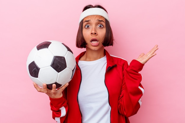 Mujer joven de raza mixta jugando al fútbol aislado sobre fondo rosa sorprendido y conmocionado.