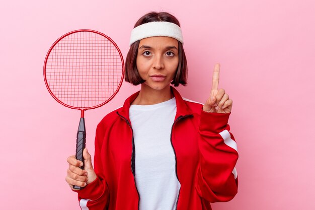 Mujer joven de raza mixta jugando bádminton aislado en la pared rosa mostrando el número uno con el dedo.