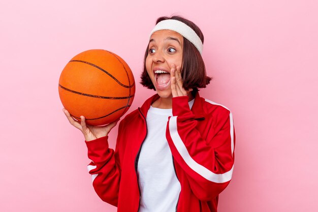 Mujer joven de raza mixta jugando baloncesto aislado en la pared rosa gritando y sosteniendo la palma cerca de la boca abierta.