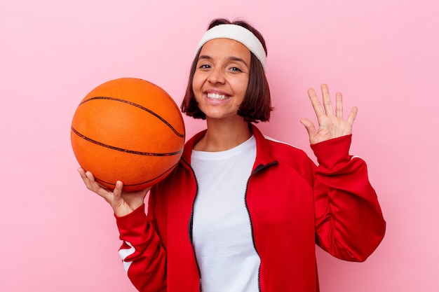 Mujer joven de raza mixta jugando baloncesto aislado en la pared rosa sonriendo alegre mostrando el número cinco con los dedos.