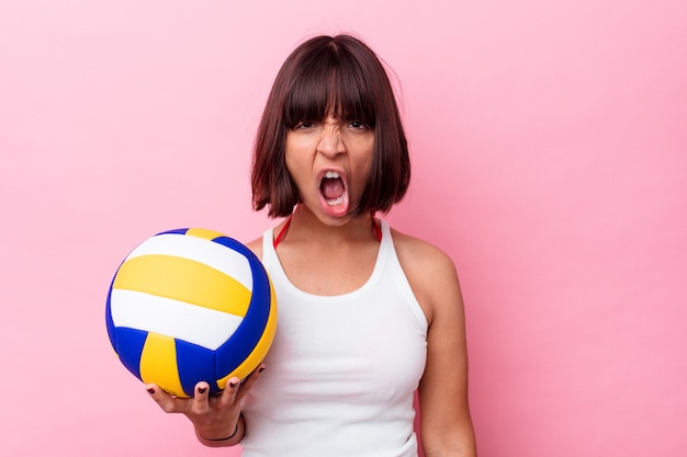 Mujer joven de raza mixta jugando voleibol aislado sobre fondo rosa gritando muy enojado y agresivo.