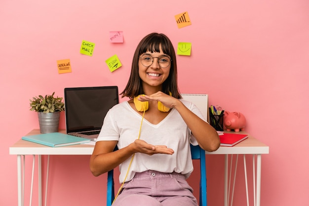 Mujer joven de raza mixta preparando un examen en la sala escuchando música aislada sobre fondo rosa sosteniendo algo con ambas manos, presentación del producto.