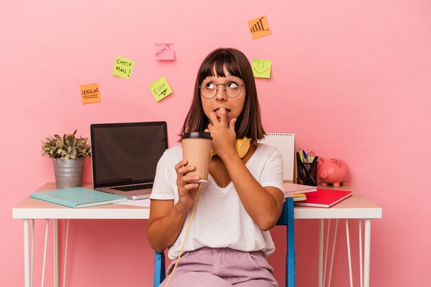 Mujer joven de raza mixta preparando un examen en la sala sosteniendo un café aislado sobre fondo rosa relajado pensando en algo mirando un espacio de copia.