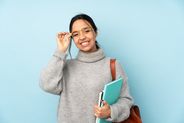 Mujer joven de raza mixta que va a la escuela en la pared azul con gafas y feliz