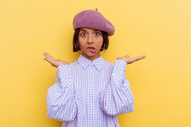 Foto mujer joven de raza mixta que llevaba una boina aislada sobre fondo amarillo confundido y dudoso encogiéndose de hombros para mantener un espacio de copia.