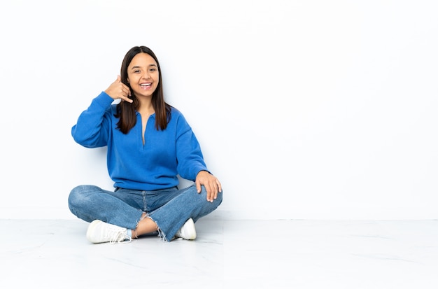 Mujer joven de raza mixta que se sienta en el piso aislado en la pared blanca que hace gesto del teléfono. Llámame señal de vuelta