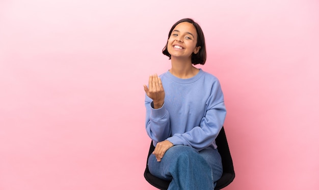 Mujer joven de raza mixta sentada en una silla aislada sobre fondo rosa invitando a venir con la mano. Feliz de que hayas venido