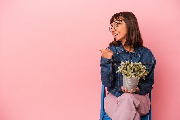 Mujer joven de raza mixta sentada en una silla sosteniendo una planta aislada en puntos de fondo rosa con el dedo pulgar, riendo y despreocupado.