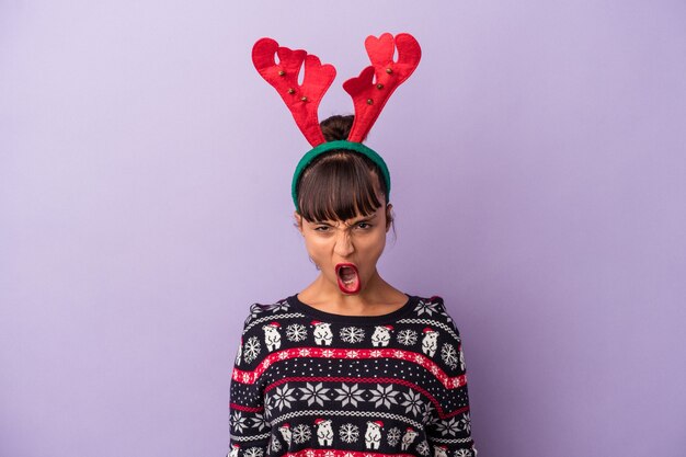 Mujer joven de raza mixta con sombrero de reno celebrando la Navidad aislado sobre fondo púrpura gritando muy enojado y agresivo.