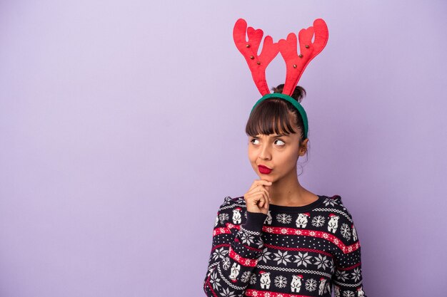 Mujer joven de raza mixta con sombrero de reno celebrando la Navidad aislado sobre fondo púrpura mirando hacia los lados con expresión dudosa y escéptica.