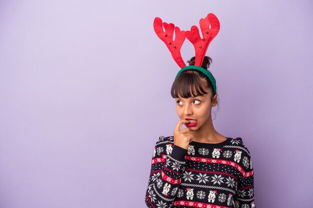 Mujer joven de raza mixta con sombrero de reno celebrando la Navidad aislado sobre fondo púrpura relajado pensando en algo mirando un espacio de copia.