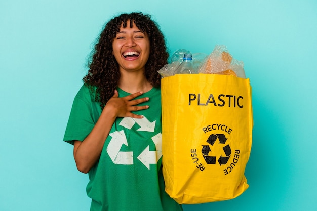 Mujer joven de raza mixta sosteniendo una bolsa de plástico reciclada aislada sobre fondo azul se ríe a carcajadas manteniendo la mano en el pecho.